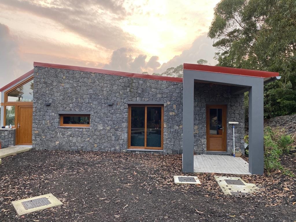 The exterior features a stone façade combined with modern design elements. Large windows allow natural light into the dwelling, while a welcoming entrance is framed by a sleek canopy. Surrounding landscaping includes natural foliage and pathways, complementing the sustainable architecture.