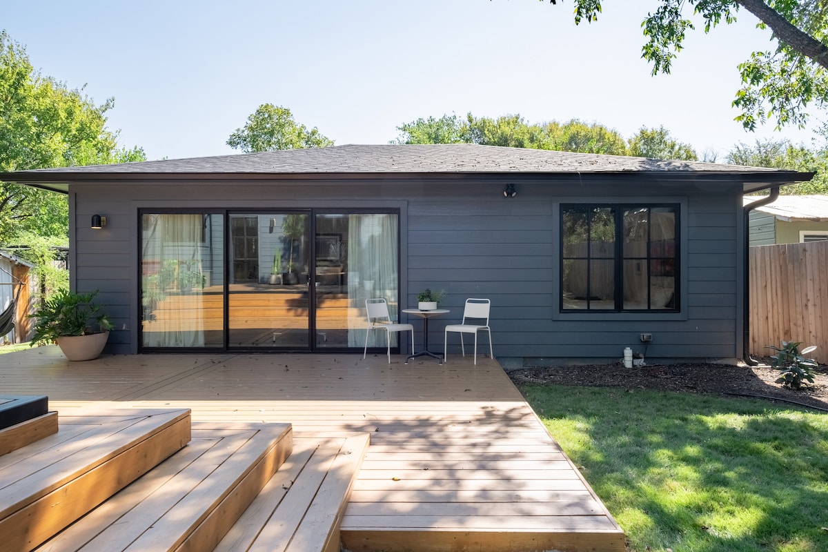 A guest house exterior is shown, featuring a spacious deck with two white chairs and a small table. Large sliding glass doors provide a view into the living space, while a well-maintained lawn and trees offer a serene setting.