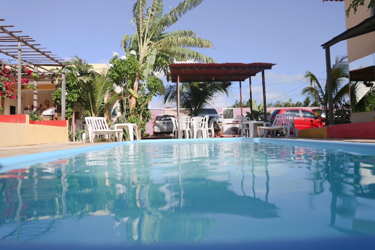A serene pool is visible in the foreground, reflecting the clear sky. Surrounding it are several white seating options and shaded areas. Palm trees and colorful plants provide a natural ambiance, while parked cars can be seen in the background, indicating accessible parking.