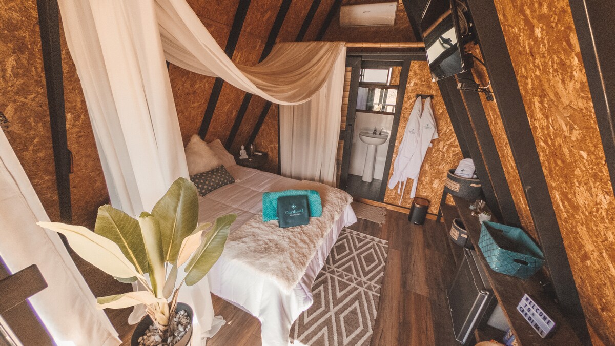 A cozy sleeping area features a spacious bed draped with soft fabrics. Natural wood walls and rich textures create a warm environment. A plush rug and a houseplant add to the inviting nature. An en-suite bathroom is visible through a nearby doorway.