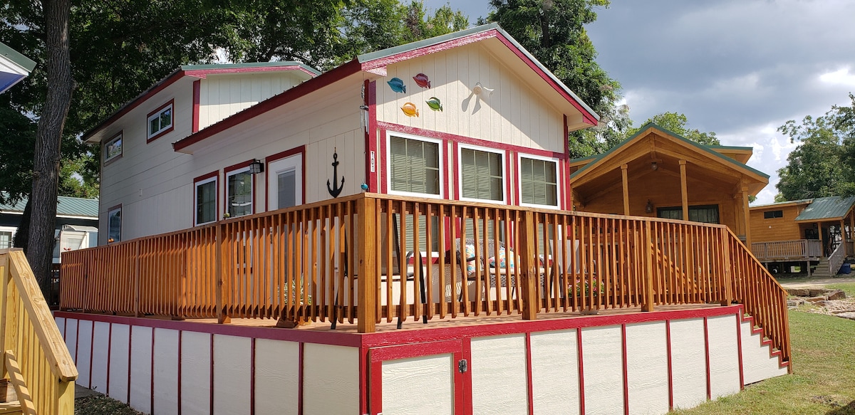 A charming cabin-style home features a large wooden deck surrounded by a railing. Colorful decorative accents are positioned above the windows, while a second smaller structure is visible in the background. Green trees lend a natural backdrop to the inviting exterior.