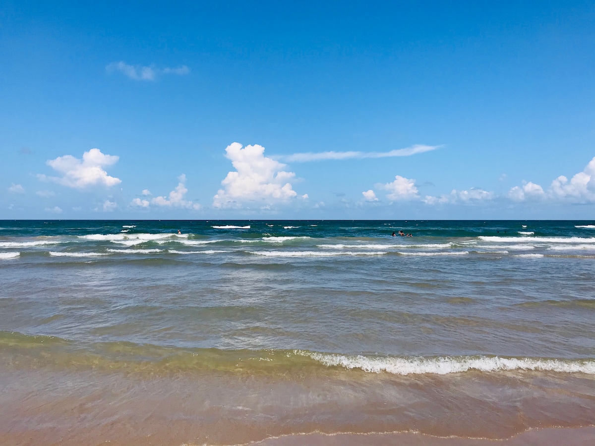 The image displays a serene beach scene with gentle waves lapping at the shore. The ocean stretches towards the horizon under a clear blue sky dotted with fluffy white clouds.