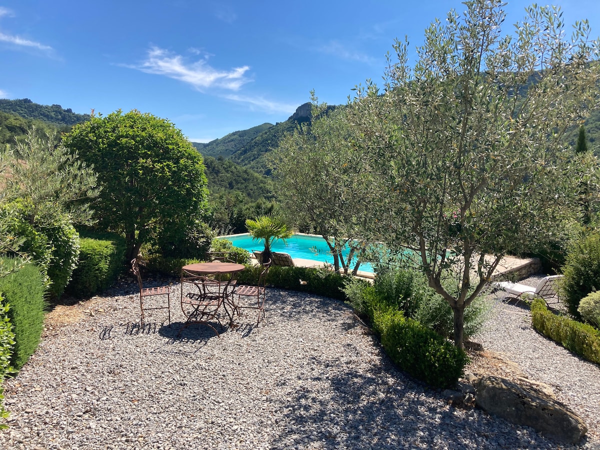 A landscaped garden features a round stone table surrounded by four chairs, overlooking a serene swimming pool. Olive trees and lush greenery frame the area, enhancing the natural beauty of the scenic hillside in the background under a clear blue sky.