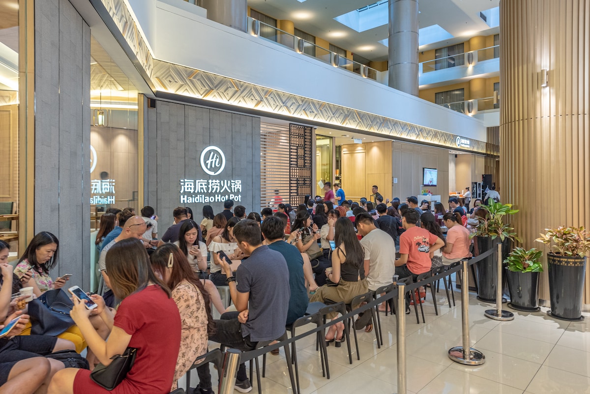 A busy interior space is filled with a crowd seated on sleek black benches. The modern decor features wooden panels and plants, while a sign indicates the presence of a restaurant. Soft lighting highlights the activity in this vibrant gathering area.