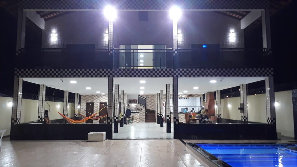The exterior view of the property showcases a well-lit, modern design with a spacious outdoor area. A swimming pool reflects the surrounding lights, and hammocks are visible in a covered lounge space. Indoor areas are open, revealing a kitchen and dining area.