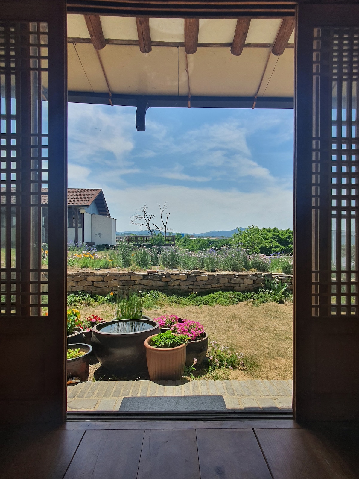 A view from the entrance reveals an outdoor space, featuring a garden with vibrant flowers in various planters. A stone wall and green trees create a serene backdrop under a clear blue sky, enhancing the tranquil environment.