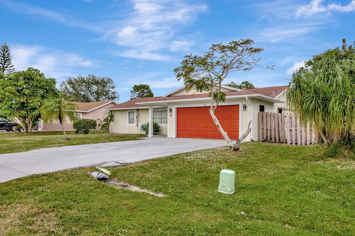 A charming single-story home is set against a bright blue sky. The residence features a welcoming orange garage door, a well-maintained lawn, and surrounding green landscaping. A concrete driveway leads to the entrance, with a fence partially visible, enhancing the property's sense of privacy.