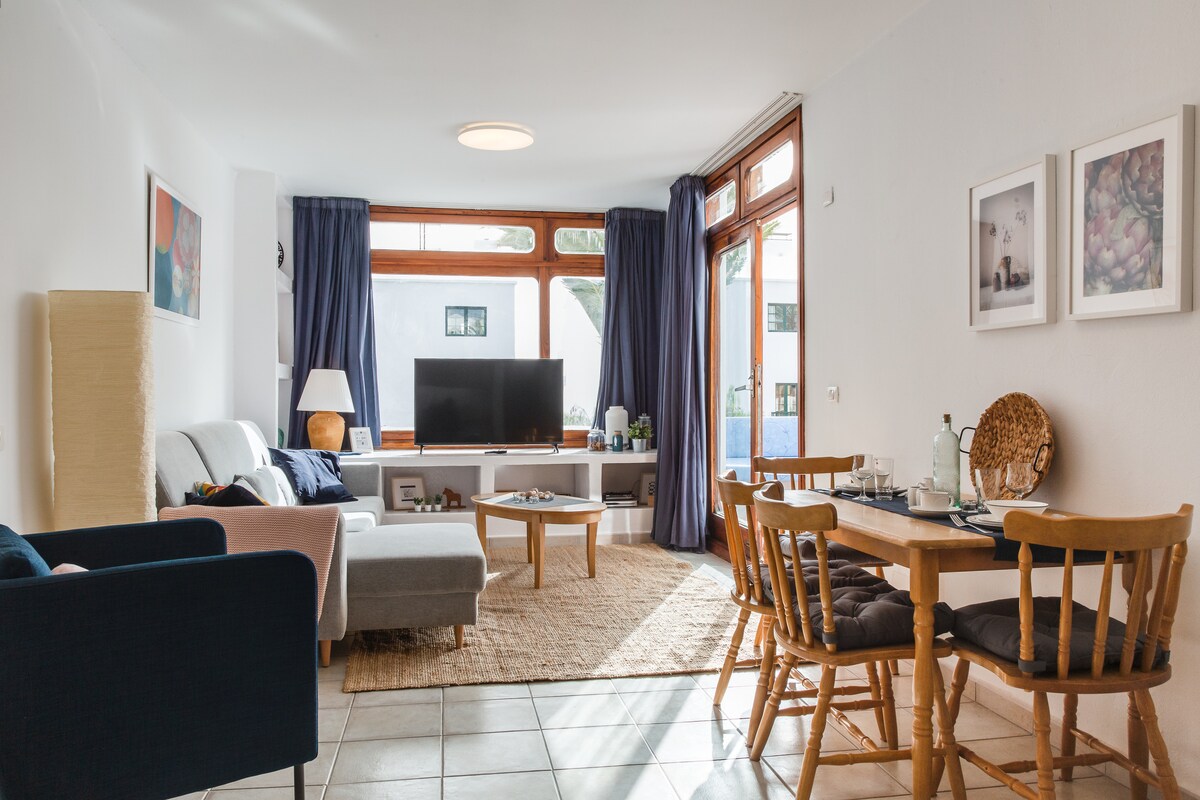 A well-lit living area is featured, showcasing a large smart TV facing a comfortable seating arrangement. Two chairs and a wooden dining table for four enhance the space. Natural light pours through large windows, framed by dark curtains, while a textured rug adds warmth to the tiled floor.