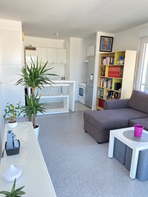 An airy living space features a comfortable grey sofa paired with a small white coffee table. A vibrant purple cup sits atop the table. Light green plants add a touch of nature, while shelves with books and decorative items create a layered backdrop.