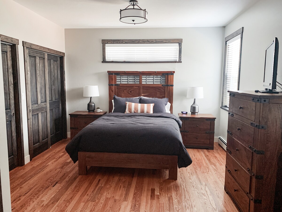 A serene bedroom is highlighted by a large wooden bed with a dark comforter. Two bedside lamps provide soft lighting, while a wooden dresser stands against the wall. Natural light filters through the window, casting gentle shadows on the warm, hardwood floor.