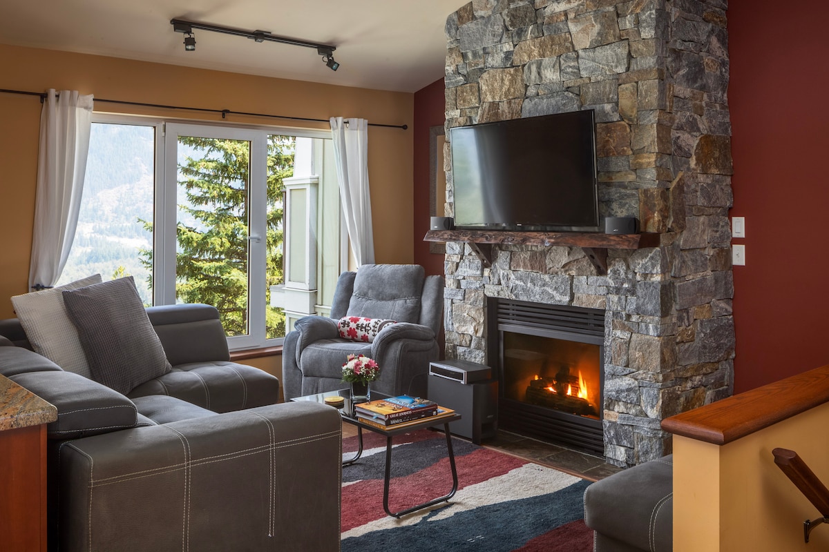 A cozy living area features a stone fireplace with a mounted flat-screen TV above it. Comfortable seating is arranged around a decorative rug. Natural light floods the space through large windows, offering expansive views of the surrounding landscape.