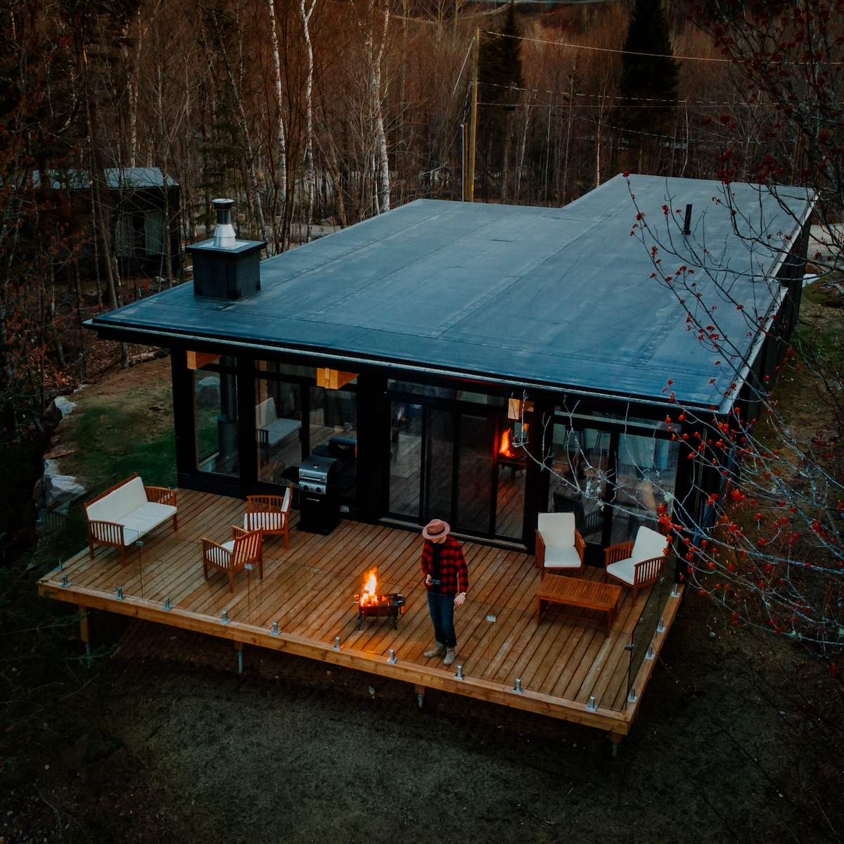 The cabin's exterior is viewed from above, showcasing a spacious wooden deck with comfortable seating and a built-in fire pit. A person stands near the fire, surrounded by a serene natural landscape with trees and a clear sky.