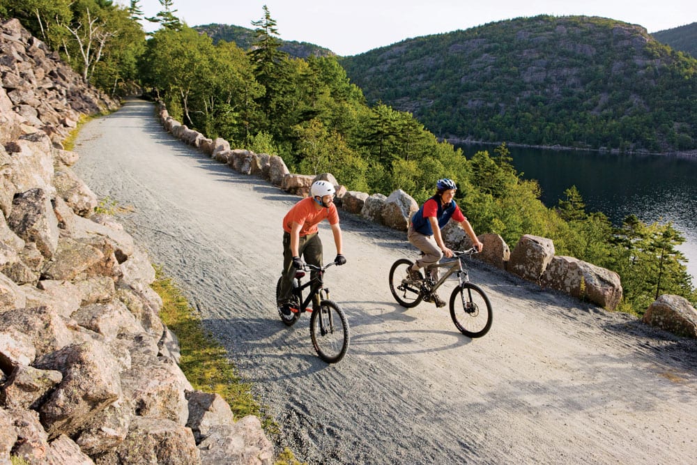 Two cyclists are riding along a gravel path bordered by large, natural stone formations. Lush greenery and a body of water are visible in the background, framed by hills. The scene suggests a peaceful outdoor setting ideal for exploration.