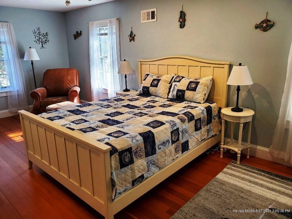 The master bedroom features a spacious bed with a patterned quilt, complemented by two bedside lamps. A cozy armchair sits in the corner, and natural light filters through sheer curtains, illuminating a hardwood floor. Nautical decorations adorn the walls, adding character to the space.
