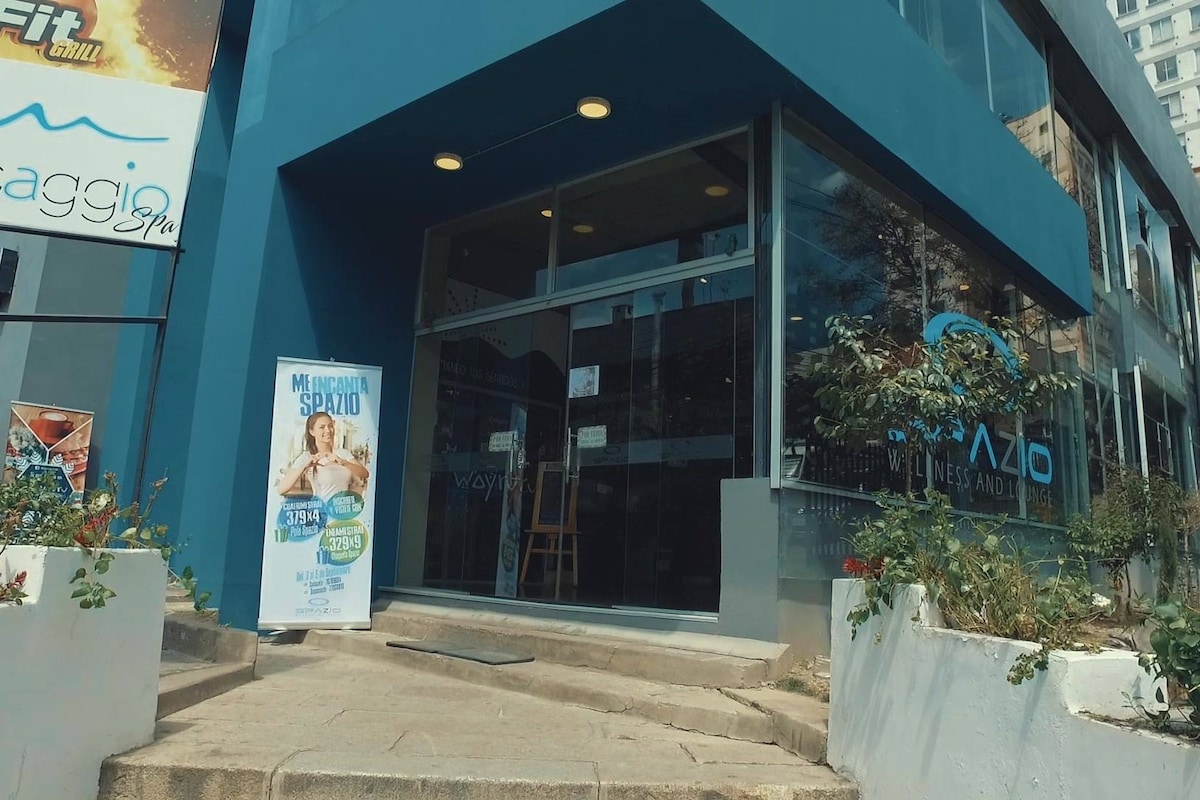 The entrance of a modern building is depicted, featuring large glass doors that provide visibility into the interior. A sign is positioned beside the door, with promotional material displayed. The path is accessible, with gentle steps leading up to the entrance, surrounded by small plants.