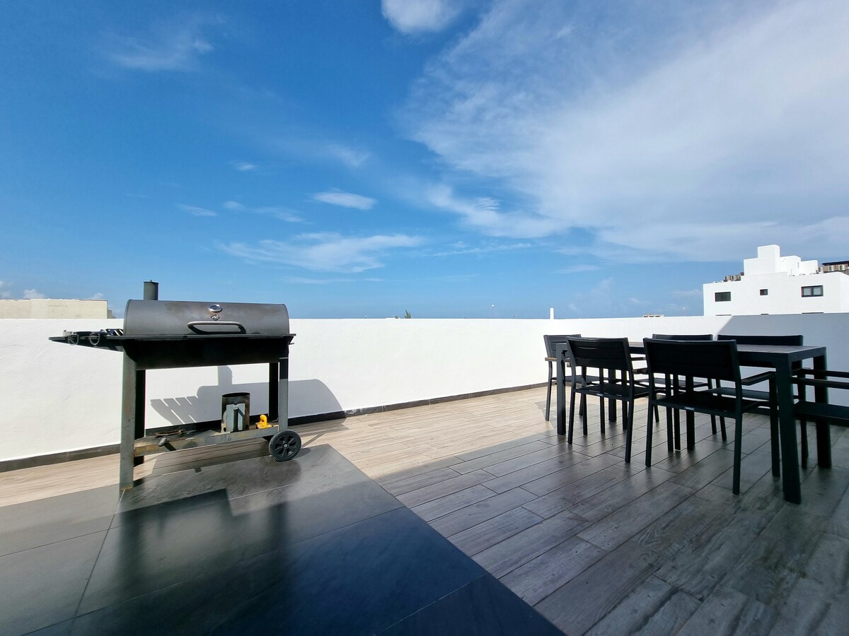 An outdoor terrace features a charcoal grill on a sturdy cart and a dining table with four chairs. The smooth flooring extends across the space, which is bathed in natural light under a clear blue sky.