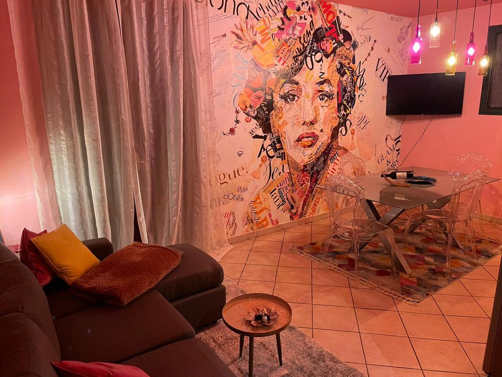 A vibrant living space features a mix of pink walls adorned with artistic patterns and a large television mounted nearby. A modern dining table is set with transparent chairs, while a plush sofa with colorful cushions invites relaxation. Soft lighting is provided by pendant lamps.