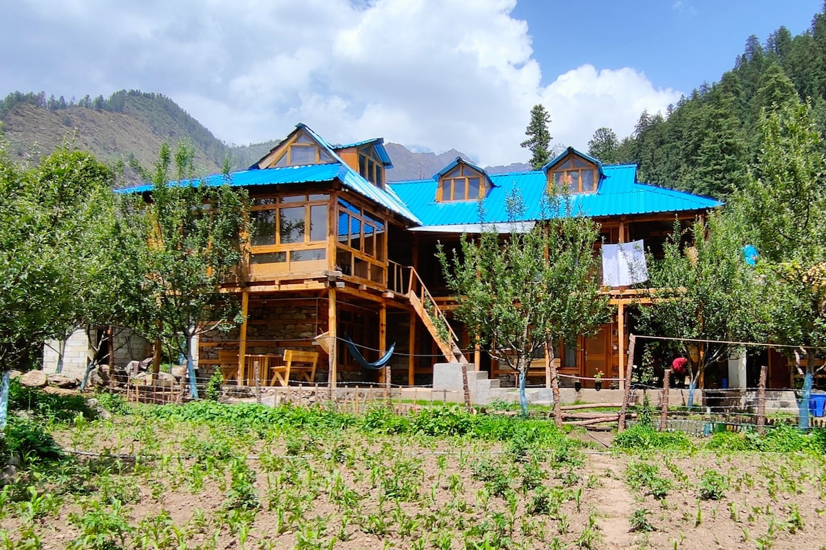 A traditional Himachali house is depicted with wooden and stone elements, surrounded by lush greenery. The two-story structure features a blue roof and large windows that invite natural light. A hammock can be seen hanging in the garden area, providing a serene spot for relaxation.