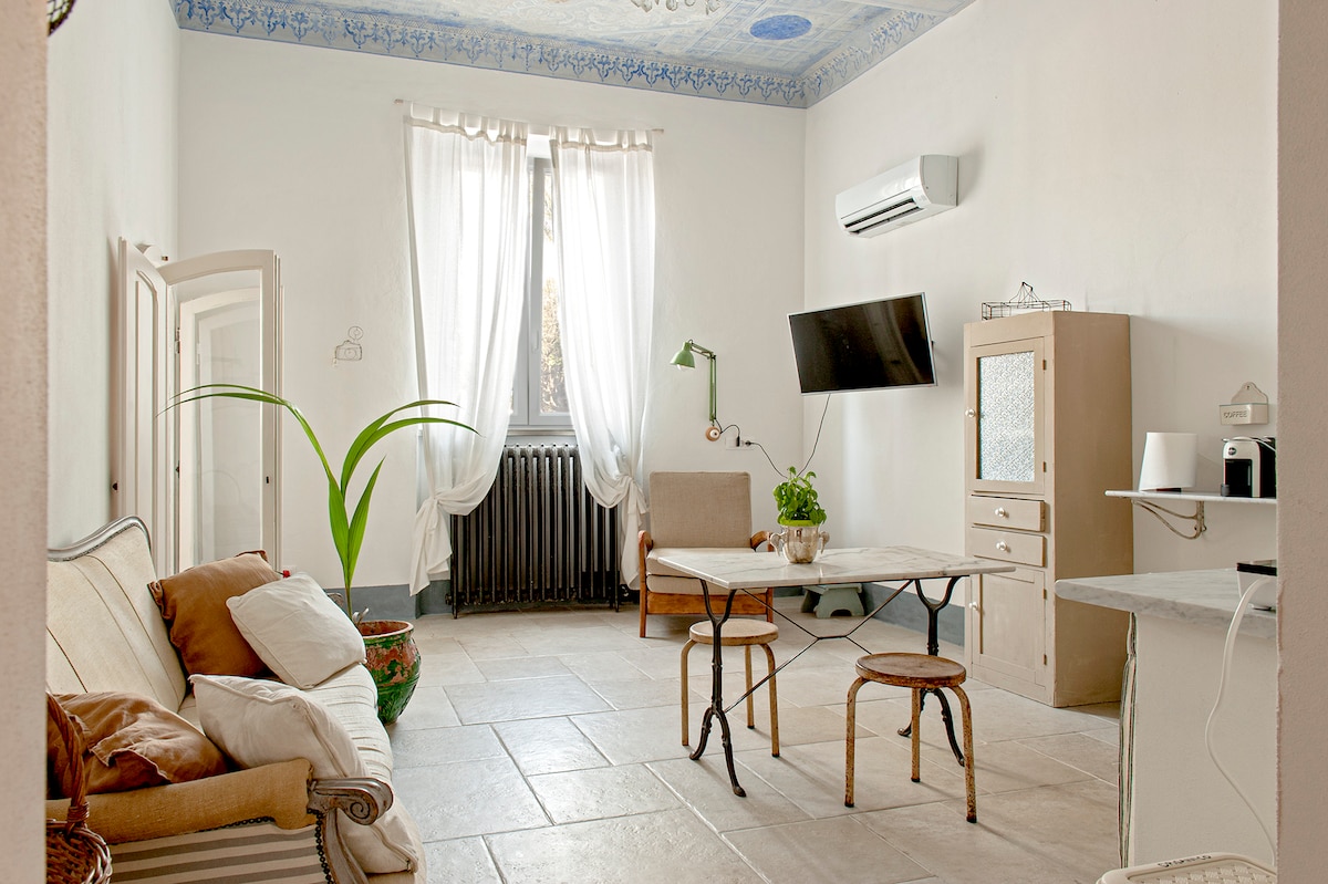 A bright and airy living space features high ceilings adorned with intricate blue details. Natural light enters through large windows dressed in sheer curtains. A cozy seating area is furnished with a sofa and armchair, complemented by a marble table and a smart TV mounted on the wall.