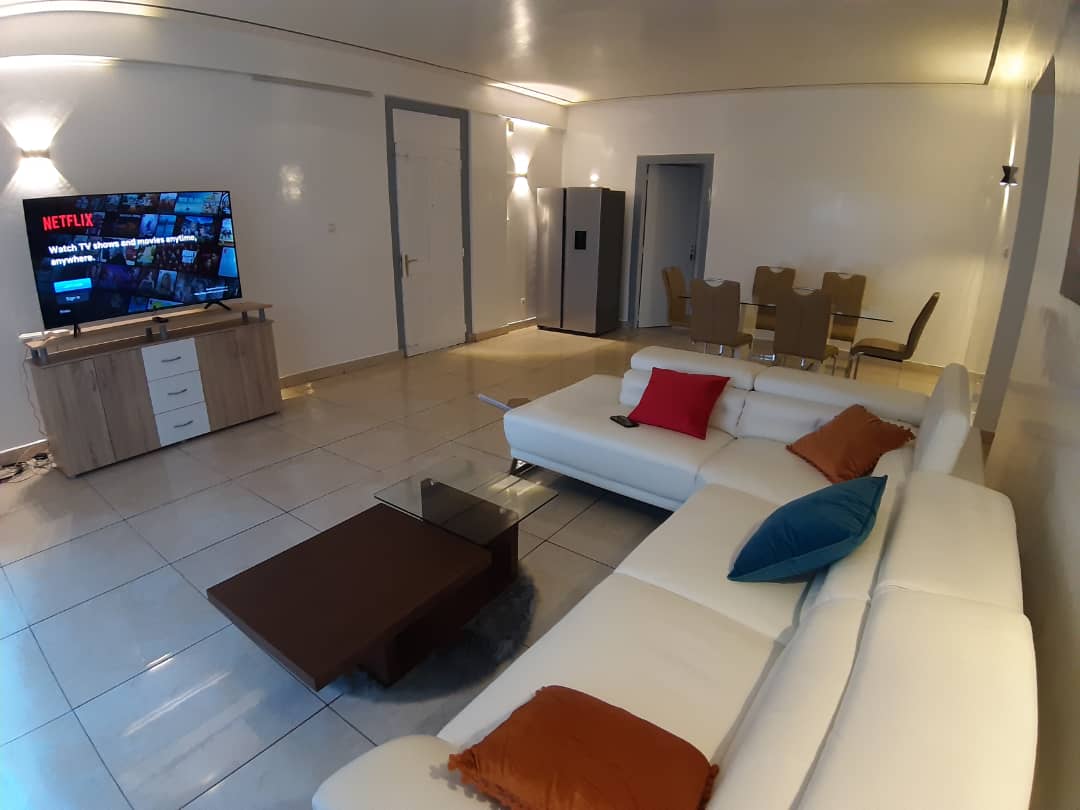 A bright living area showcases a modern white sectional sofa with colorful cushions, positioned near a coffee table. A large flat-screen TV is mounted on the wall, with a wooden entertainment unit below. The space includes a dining area with four chairs and a polished table.