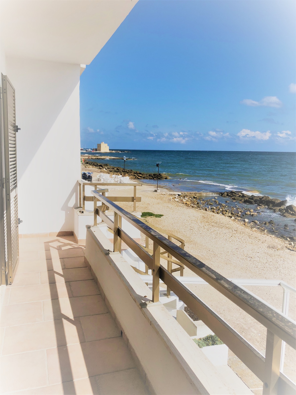 A spacious balcony is visible, offering views of the coastline and the sea. The terrace is adorned with a railing, while steps lead down to the sandy shore. The clear sky and gentle waves create a serene environment, inviting relaxation and enjoyment of the scenic surroundings.