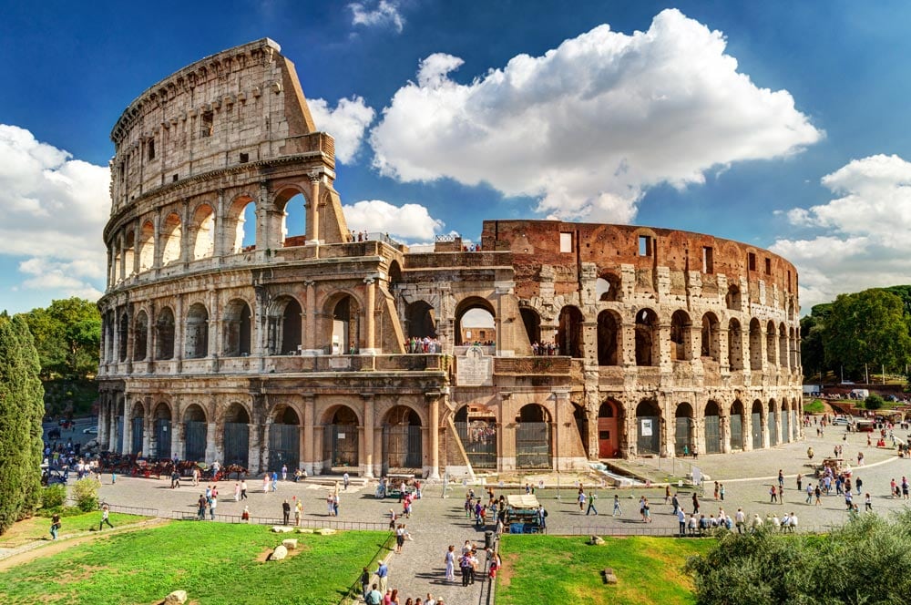 The iconic Colosseum is prominently displayed, showcasing its grand exterior with multiple arches and stonework. Visitors can be seen exploring the area around the structure, which is surrounded by lush greenery under a partly cloudy sky. The ancient monument reflects Rome's rich history.
