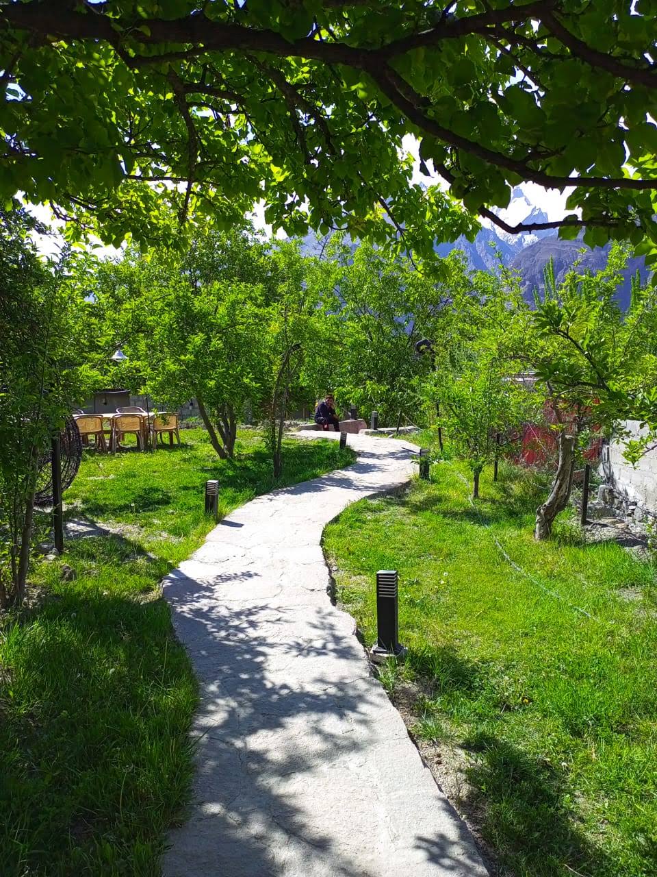 A winding stone path is visible, surrounded by lush green grass and fruit trees, leading into a vibrant garden area. Soft shadows from overhanging branches create a serene atmosphere. A small seating area with chairs is positioned in the distance, inviting relaxation amidst nature.