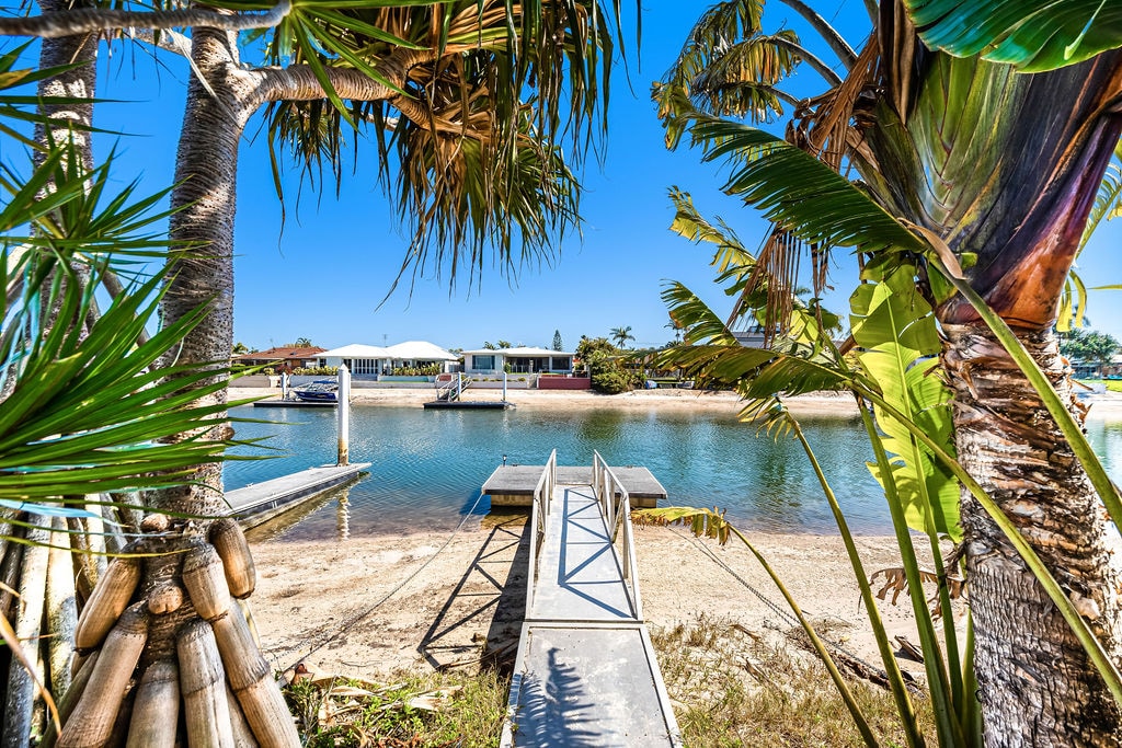 A calm waterfront view showcases a private boat ramp leading to a serene canal. Lush palm trees frame the scene, providing a tropical atmosphere. The tranquil water reflects clear blue skies, with modern homes visible in the background.