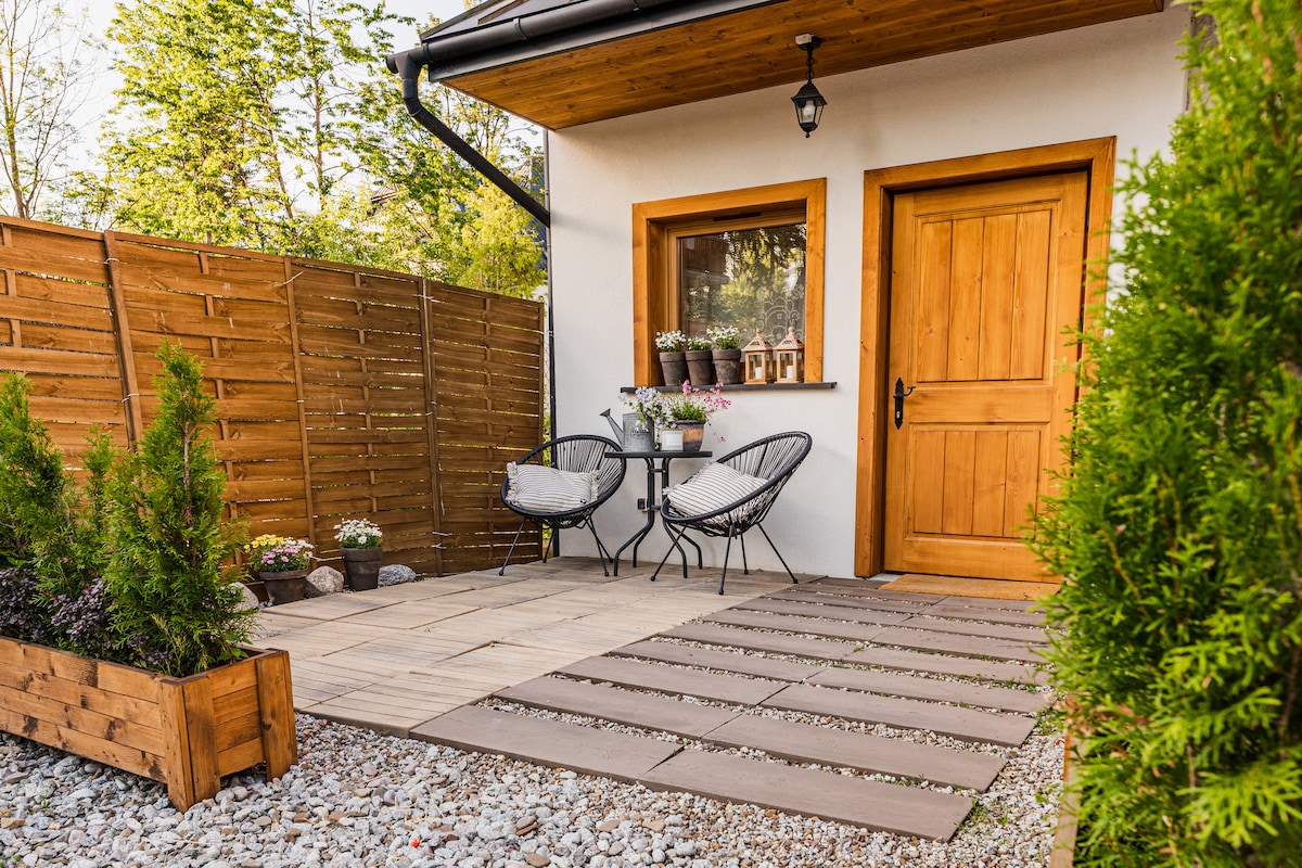 A private terrace is shown, featuring two chairs and a small table nestled against a wooden fence. Planters filled with flowers add greenery, while a wooden door with a bold frame stands as the entrance to the apartment.