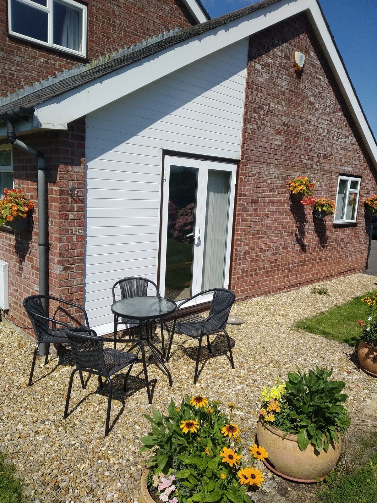 An outdoor seating area is presented with a small round table and four black chairs, surrounded by well-maintained gravel and vibrant planters. Bright sunflowers and lush greenery enhance the welcoming space, with a clear blue sky and sunlight illuminating the setting.