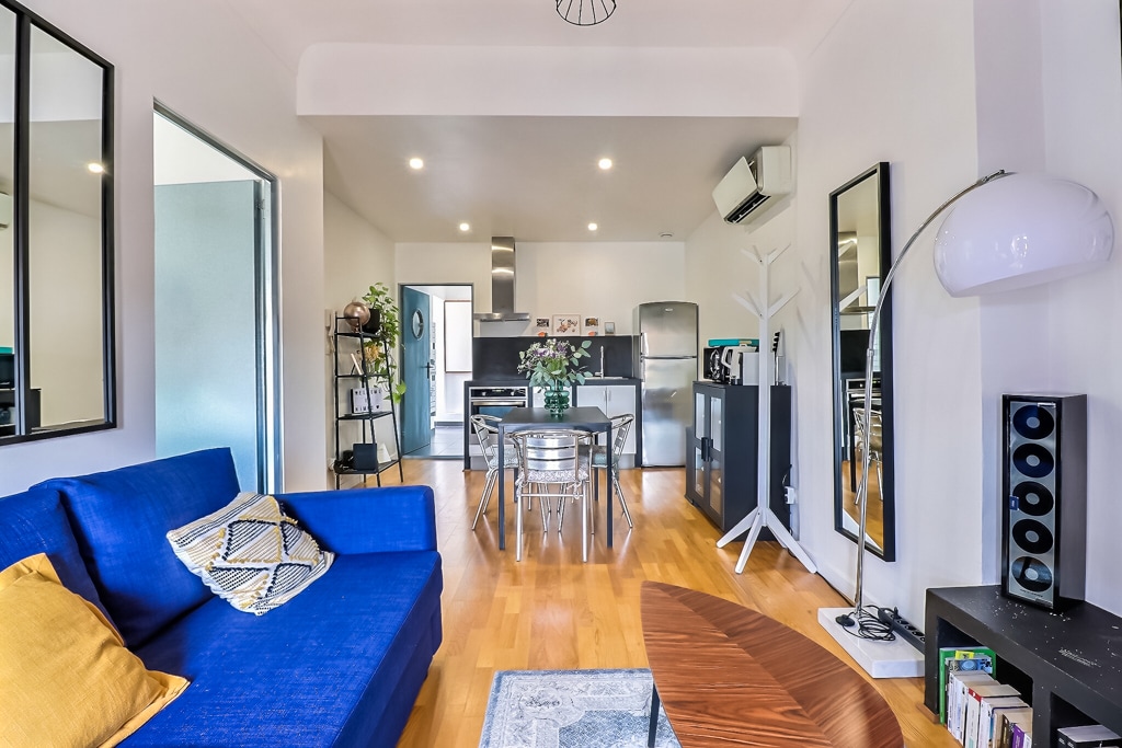 A modern living space features a vibrant blue sofa with a decorative pillow and a stylish wooden coffee table. A dining area with a round table and four chairs is visible, alongside a sleek kitchen equipped with stainless steel appliances and a decorative plant.
