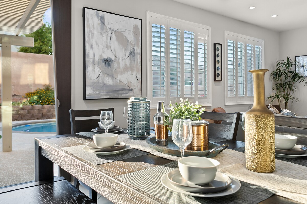 A dining area is set for a meal, featuring a long table adorned with stylish tableware. Soft natural light enters through the plantation shutters, highlighting contemporary wall art and a decorative vase. The inviting ambiance is complemented by a view of the private backyard.