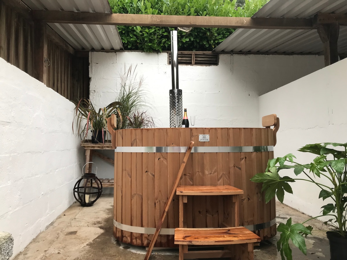 A wood-fired hot tub is situated in a private, sheltered area, surrounded by greenery. The circular tub features a wooden exterior and a metal chimney. A wooden step leads up to the edge with two small wooden tables beside it, adding a natural touch to the space.