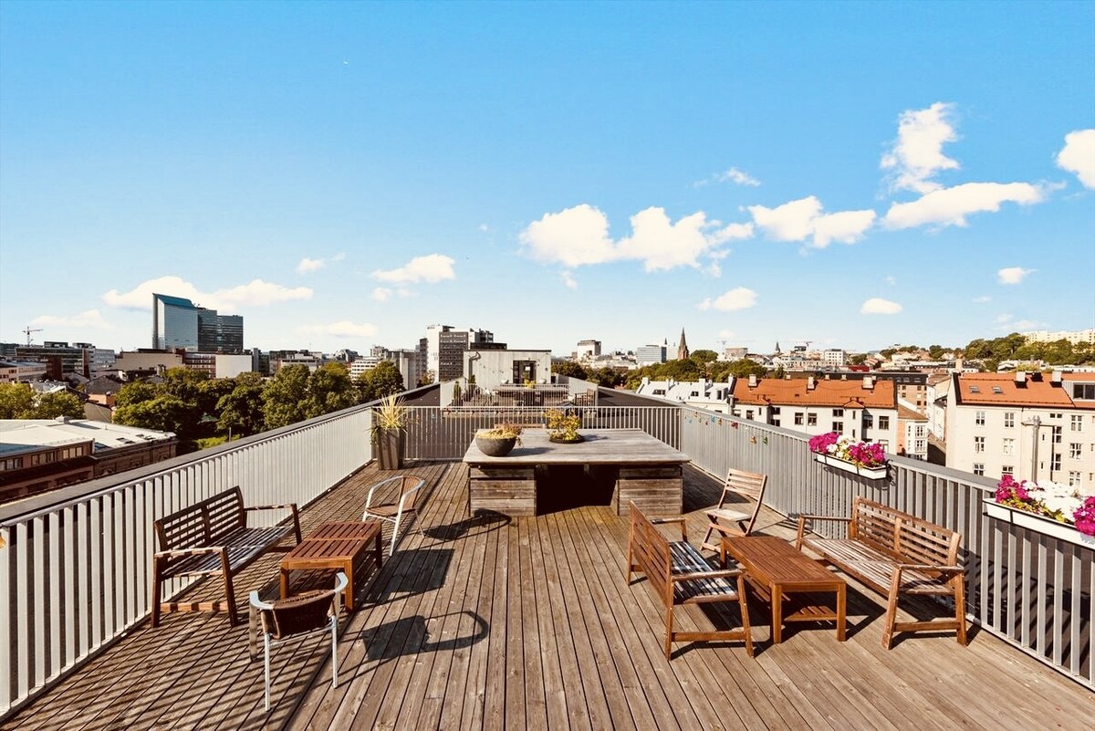 A rooftop balcony is presented with wooden decking, featuring an arrangement of several outdoor seating options. Planters with greenery add a touch of nature, while panoramic views of the city skyline are visible against a clear blue sky.