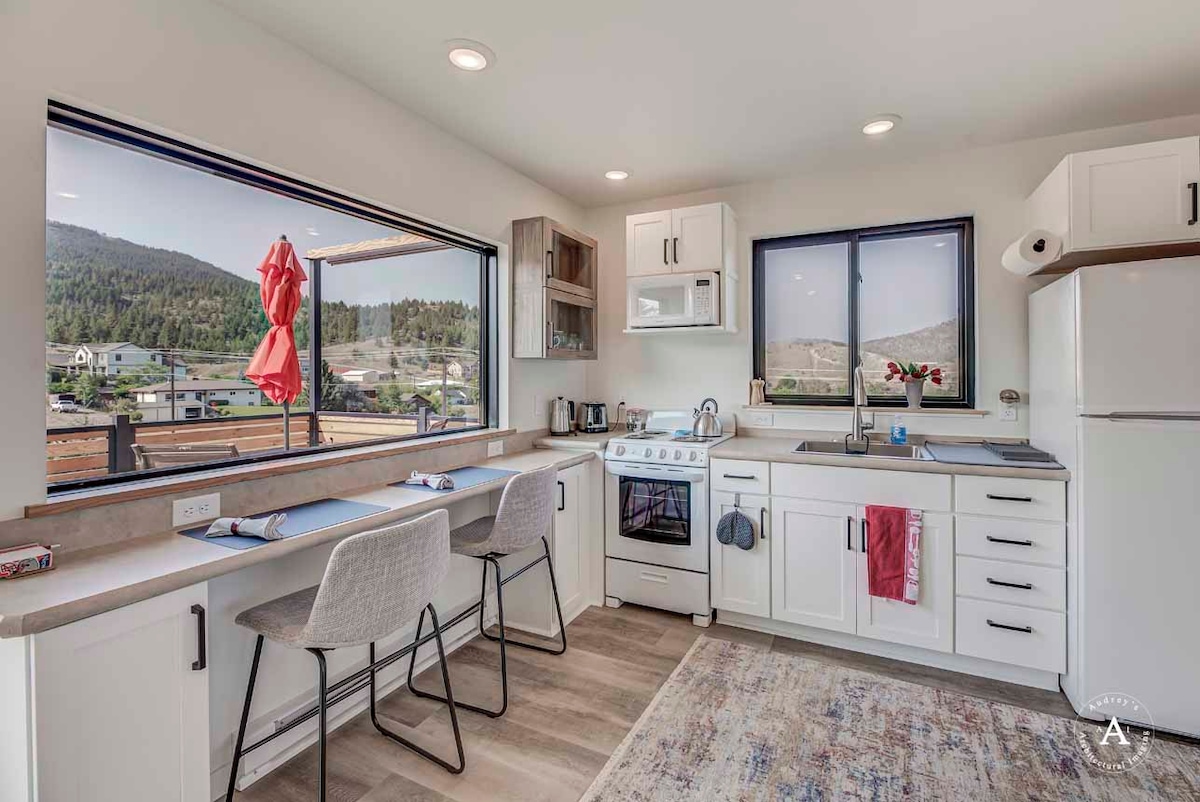A modern kitchen is illuminated by natural light from large windows. Features include a full range, microwave, and refrigerator. Counter seating with two stools is available, alongside contemporary cabinetry. Towels and an apartment-sized coffee maker are visible, with a picturesque mountain view beyond the windows.