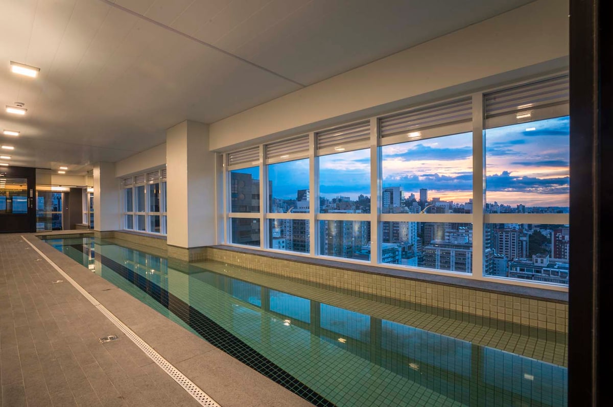 A spacious indoor pool area is illuminated by soft lighting, featuring large windows that provide a wide view of the city skyline. The pool is lined with tiles, and the sleek surrounding walls enhance the contemporary design.