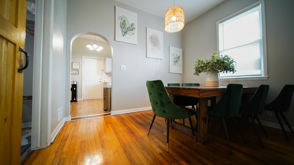 A dining area features a wooden table surrounded by green chairs. Natural light enters through a window, highlighting the polished wood floor. Artwork is displayed on the walls, and an archway leads toward the kitchen.