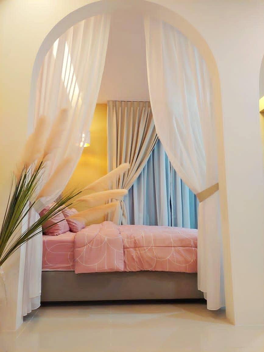 A cozy bedroom is highlighted by a plush, light pink bed, softly draped with sheer white curtains. Natural light filters through the large window, illuminating the space. A touch of greenery is presented with decorative plants beside the bed.