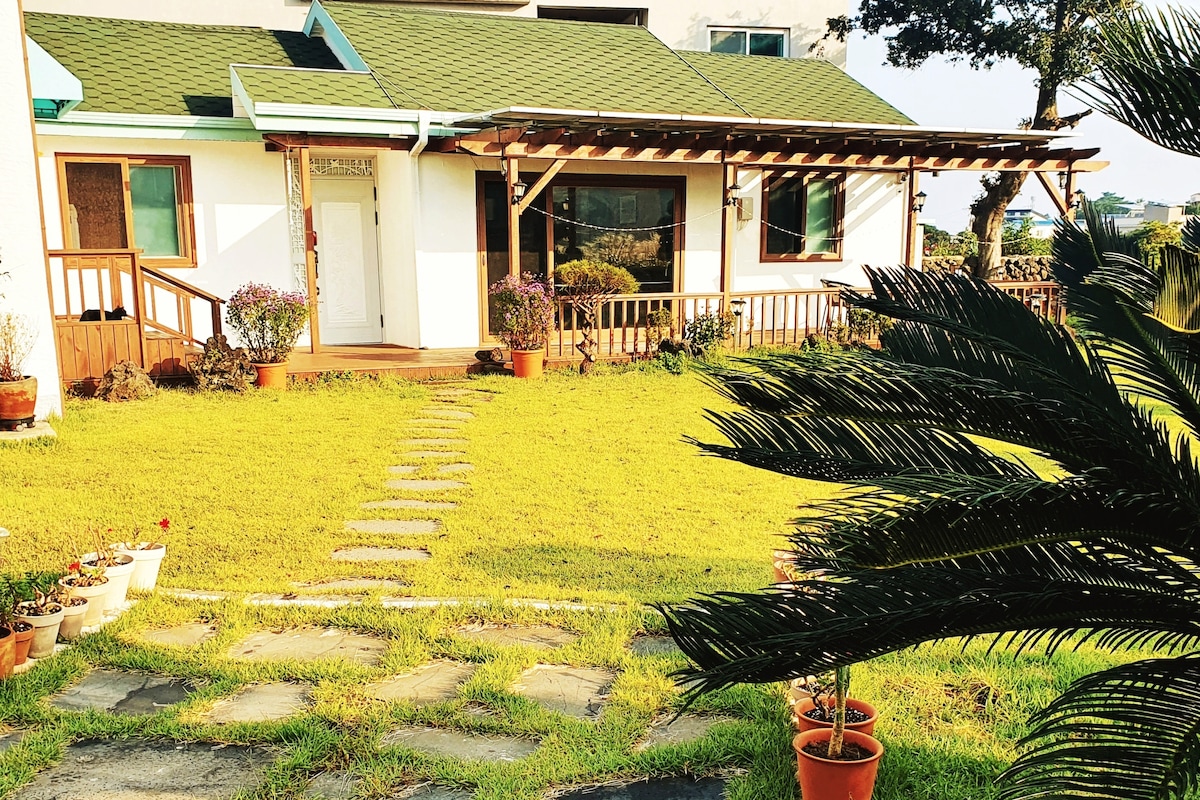 A spacious yard is presented with lush green grass and stone pathways leading to the entrance of the house. Flower pots are arranged along the path, and a wooden deck offers a cozy seating area outside. The house features large windows and a sloped roof.