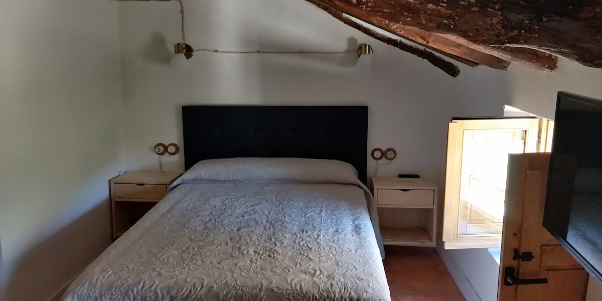A spacious sleeping area features a dark upholstered headboard and a light-colored quilt. Nightstands with circular lamps stand beside the bed. A window invites natural light, while rustic wooden beams accentuate the high ceiling.