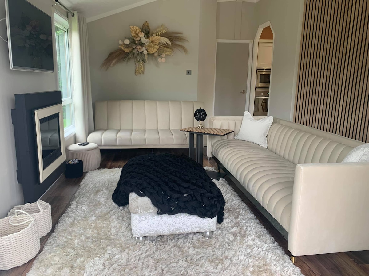 A modern living area features a light-colored, L-shaped sofa paired with a chic accent chair. A textured rug covers the floor, while a small coffee table sits in the center. Natural light is allowed in through large windows, creating a soft ambiance.