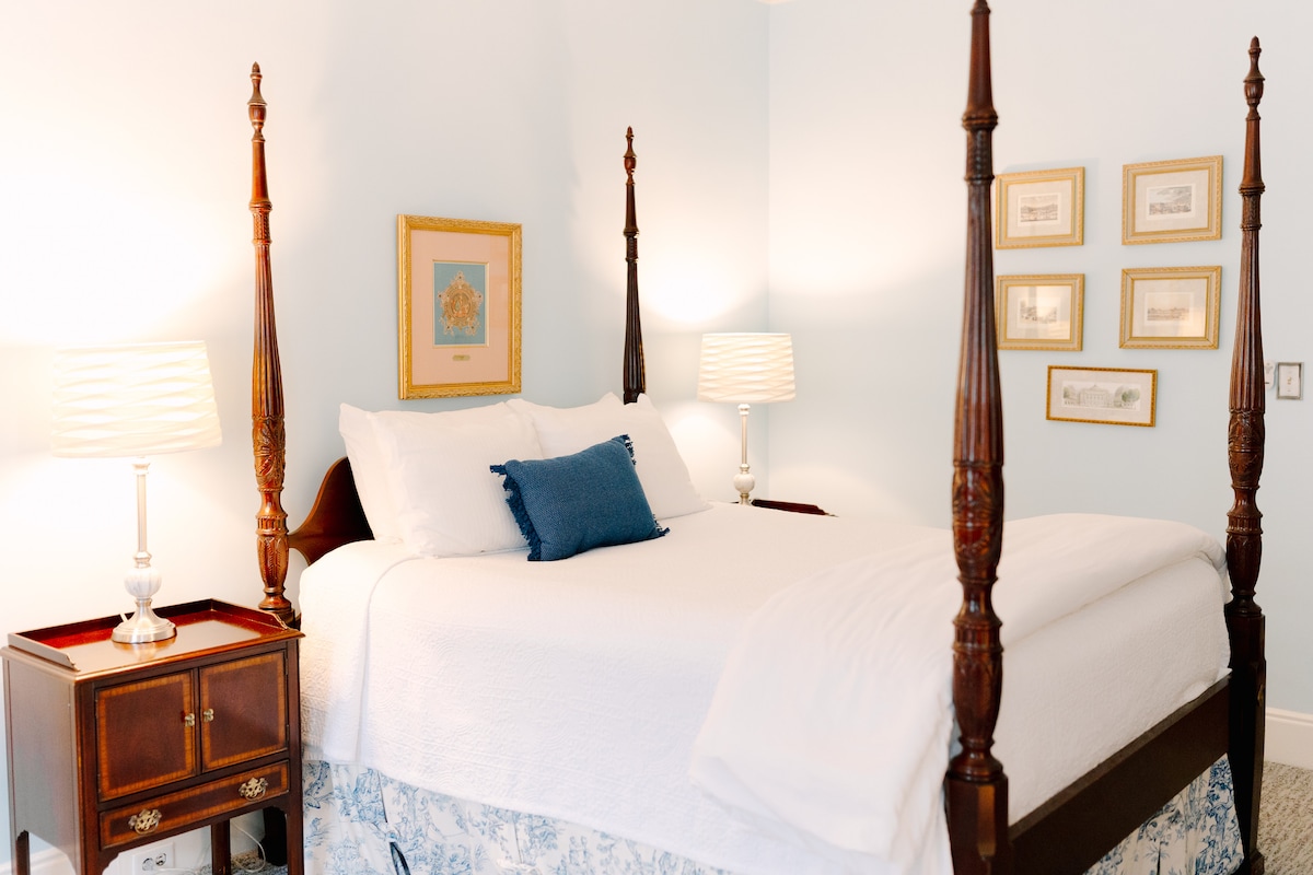 A queen-sized canopy bed is centered in the room, adorned with white linens and a blue decorative pillow. Two bedside lamps provide soft illumination, while framed artwork hangs on the pale blue walls, adding a touch of elegance to the serene environment.