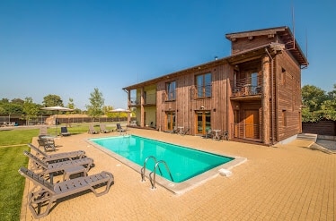 A modern wooden structure offers a spacious outdoor area featuring a sparkling swimming pool surrounded by lounge chairs. Lush greenery is visible in the background, enhancing the sense of tranquility. The building showcases large windows and multiple outdoor seating areas for relaxation.