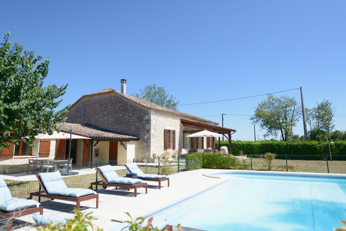 The spacious farmhouse is framed by a well-maintained garden and a swimming pool. Sun loungers are positioned beside the pool, inviting relaxation under a clear blue sky. The house features a combination of stone and wooden elements, and shaded seating is available nearby.