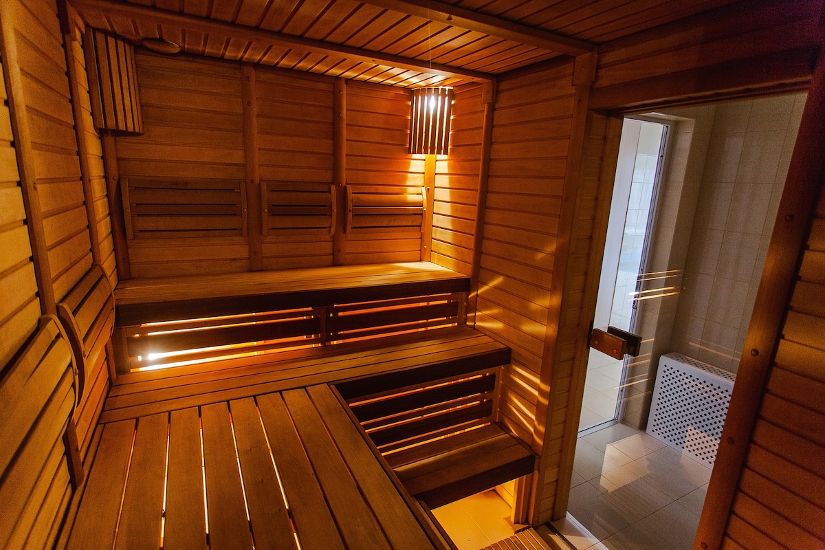A wooden sauna is presented with warm lighting highlighting the natural grains of the wood. Wooden benches create seating along the walls, while a doorway leads to an adjacent shower area. The overall design emphasizes a serene and calming atmosphere.