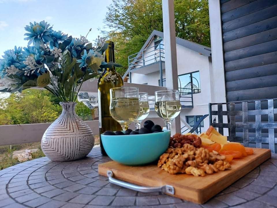 A table set for relaxation features a wooden platter with an assortment of cheese, nuts, olives, and dried fruit. Two glasses of white wine are positioned alongside, with a vase of blue flowers providing a colorful accent. A modern house is visible in the background.