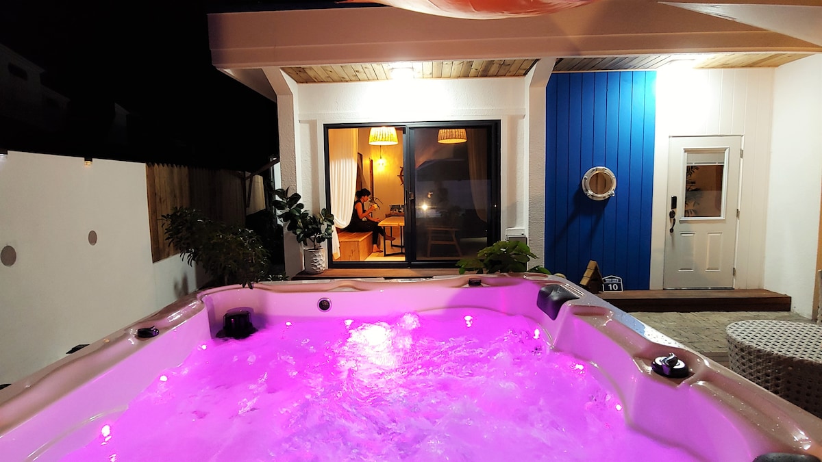 An outdoor hot tub is illuminated with soft pink lights, creating a warm atmosphere. Behind it, large glass doors lead into a cozy indoor space, where pendant lights hang and a plant adds a touch of greenery.