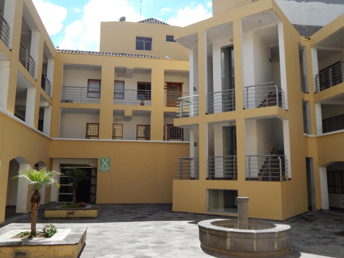 An open courtyard features a central stone fountain, surrounded by modern buildings with multiple balconies and staircases. Light-colored walls are complemented by tropical plants, creating a welcoming environment. Ample natural light filters into the space, contributing to an airy atmosphere.