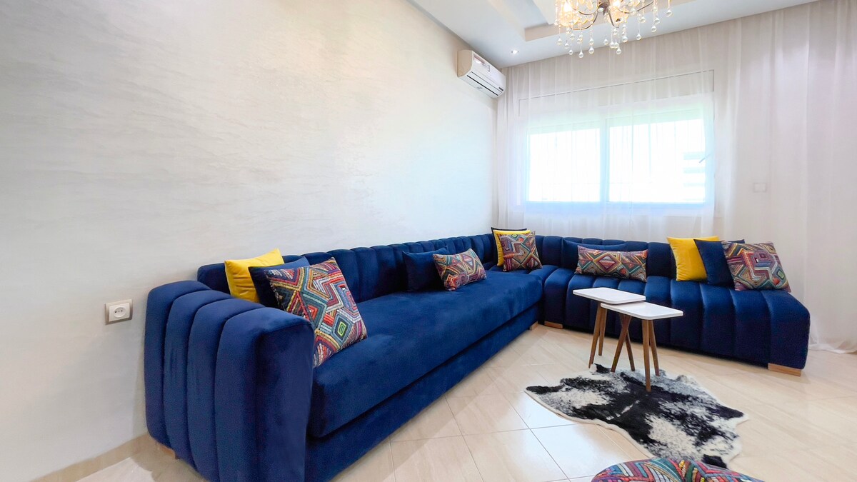 A spacious living area is furnished with a large, dark blue sectional sofa adorned with colorful patterned cushions. A small white side table is positioned in front of the sofa, complemented by a black and white rug on the tiled floor. Natural light filters in through large windows with sheer curtains.