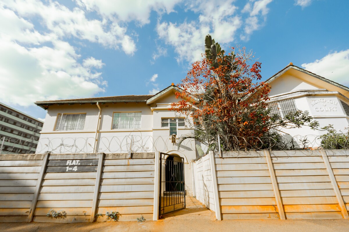 The exterior of a renovated flat is displayed, featuring a white façade and a welcoming entrance. A vibrant tree with red blossoms is visible in the foreground, while a fence surrounds the property, enhancing privacy. A clear blue sky can be seen above.